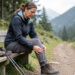 Une femme assise sur un banc de sentier tient doucement son genou douloureux après une randonnée, avec ses chaussures de marche et ses bâtons à proximité.
