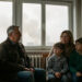 Une famille composée d’un homme, d’une femme et de deux enfants est réunie dans un logement aux fenêtres fermées, tandis qu’un panache de fumée lointain voile le ciel extérieur.