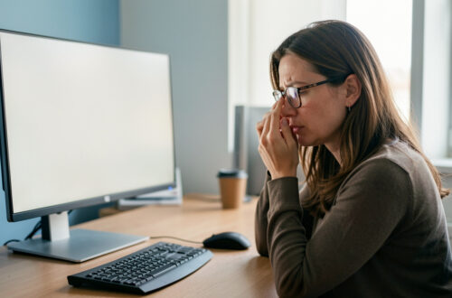 Une femme portant des lunettes se repose devant un poste informatique ergonomique et ajuste son écran dans un bureau lumineux.