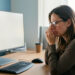 Une femme portant des lunettes se repose devant un poste informatique ergonomique et ajuste son écran dans un bureau lumineux.