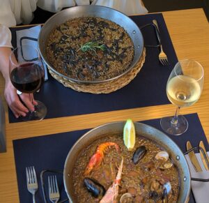 Paella garnie de fruits de mer servie avec des verres de vin blanc sur une table.
